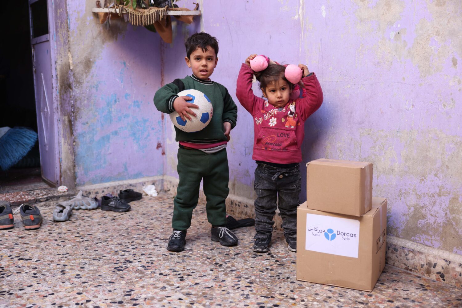 Jongetje met een bal links. Rechts een meisje wat speelt met haar haren. Daarnaast een voedselpakket van Dorcas met een dorcas logo van Dorcas Syrië