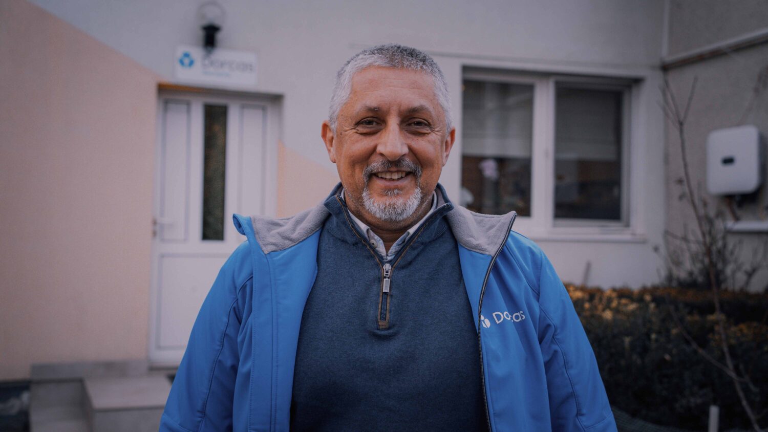 Man met grijs haar en baard, lachend in voortuin van woning, gekleed in blauwe werkjas en colbert, ambassadeur van Dorcas.