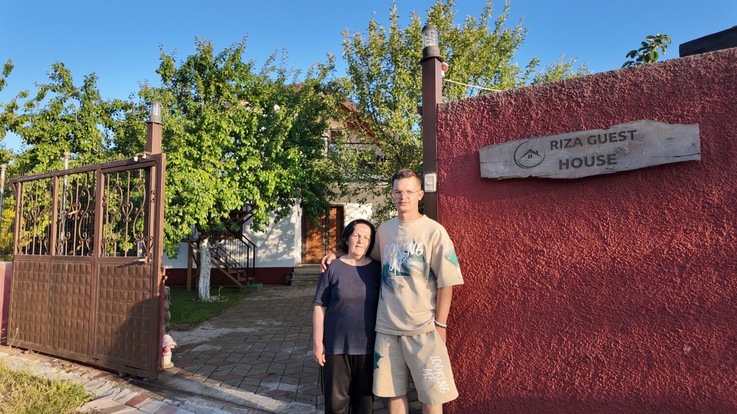 Een foto van een vrouw en een jonge man voor de ingang van Riza Guest House, omgeven door groene bomen en een heldere blauwe lucht.