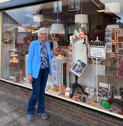 Gezellige vrouw voor winkelraam met handgemaakte handwerken, breiwerk en decoraties, winkel van Dorcas met open sign in het raam.