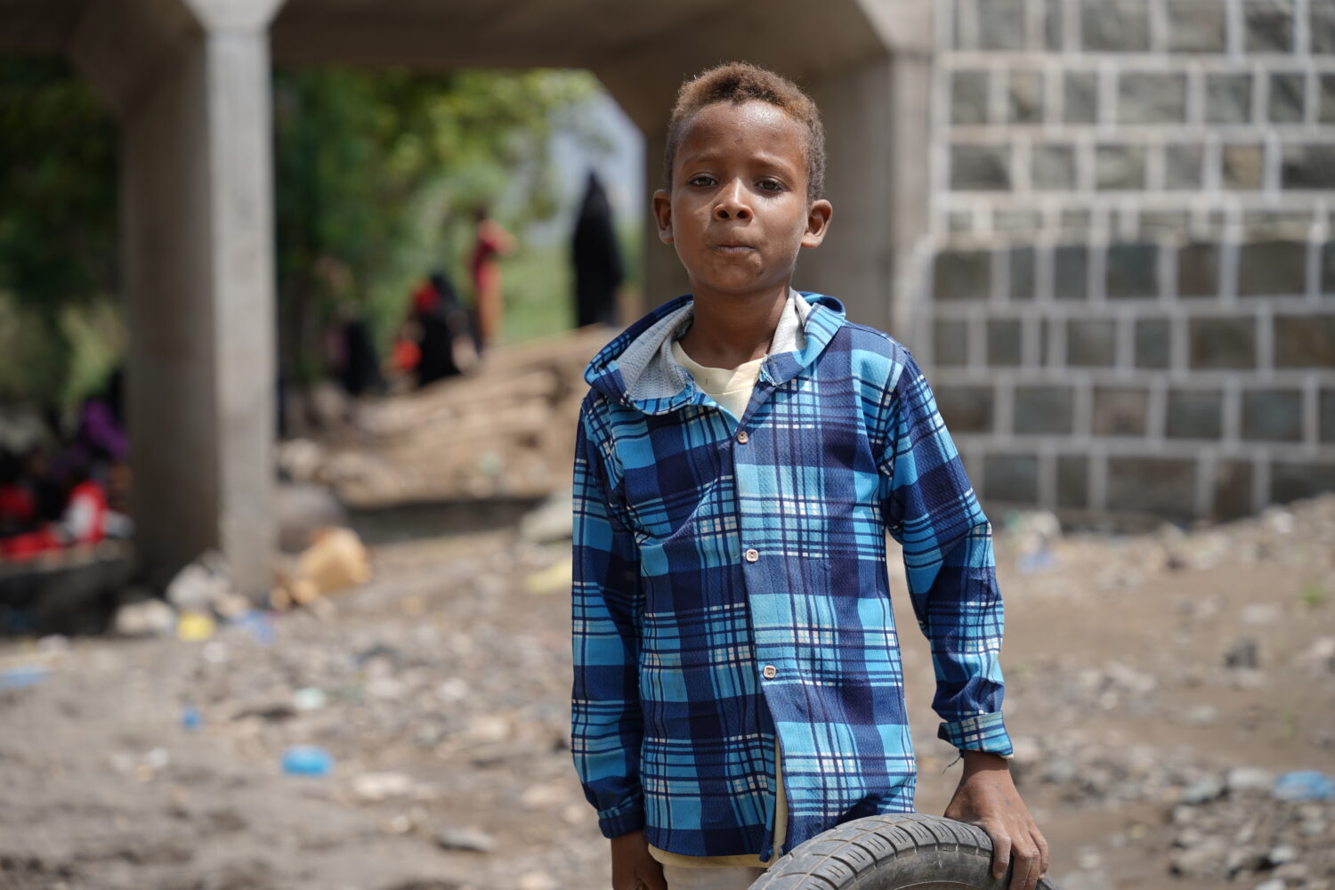 Kind met een blauwe geruite jas die een autoband vasthoudt naast een brug.