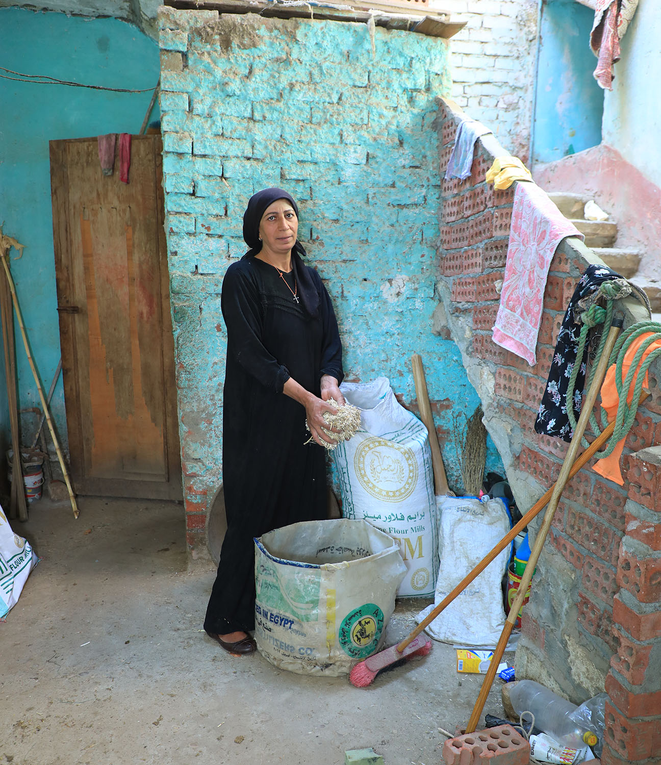 Een vrouw in een zwarte jurk en hoofddoek die op een bouwplaats staat, omgeven door bouwmaterialen en gereedschap.