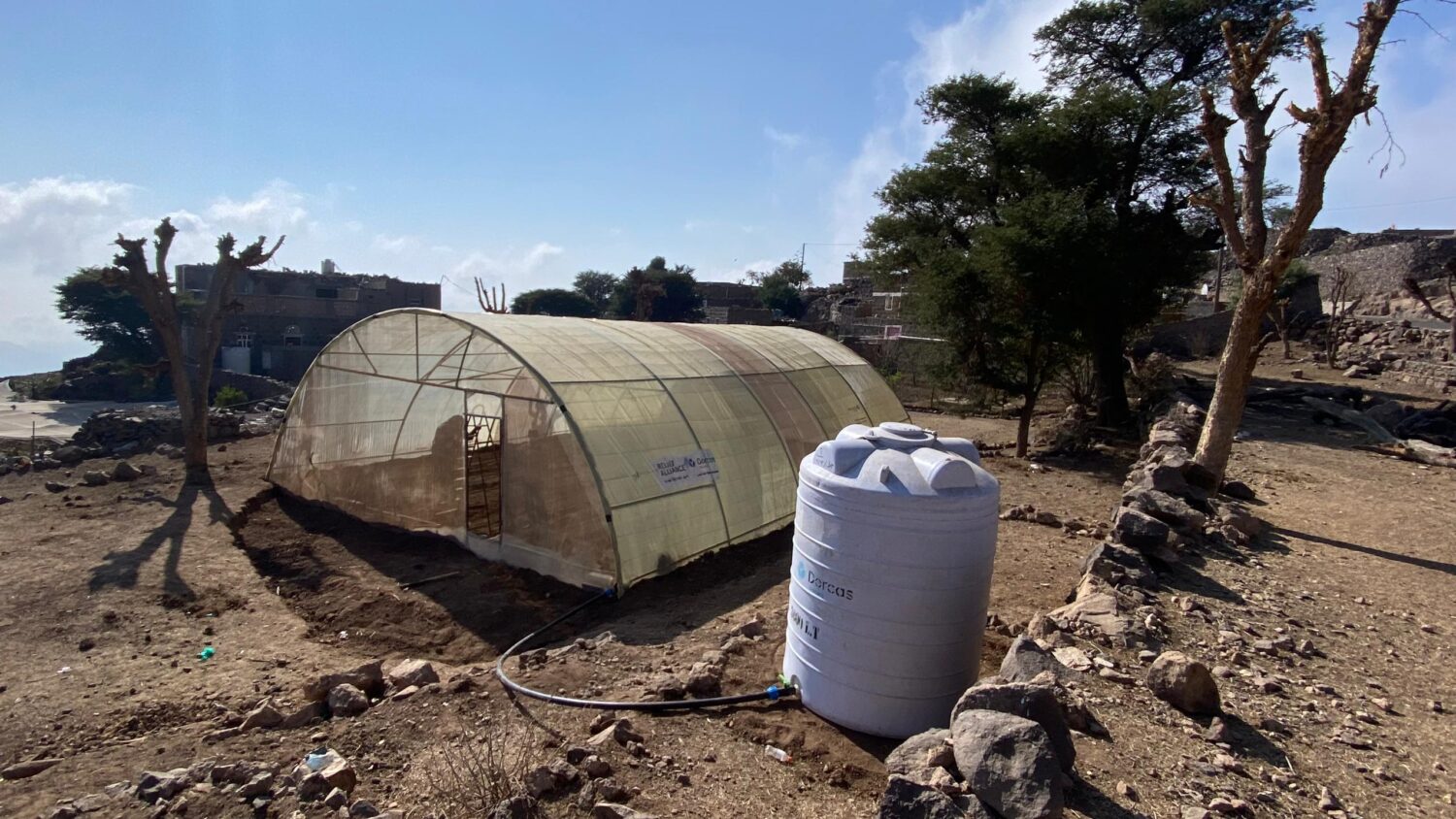Kleine kas met uitstalraam, geïnstalleerd op een drooge, steenrijke grond met stenen en bomen op de achtergrond, voor duurzame landbouw of tuinprojecten in Afrika.