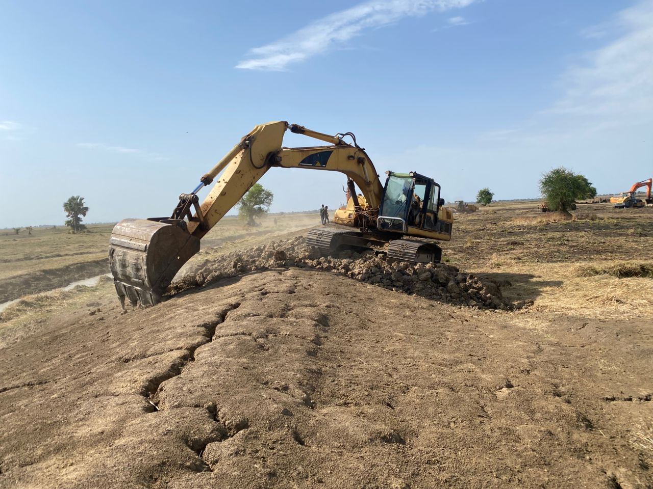 Graafmachine graaft de aarde op een open veld, duidelijk voor landbouwwerkzaamheden of infrastructurele projecten, met een heldere blauwe lucht op de achtergrond.