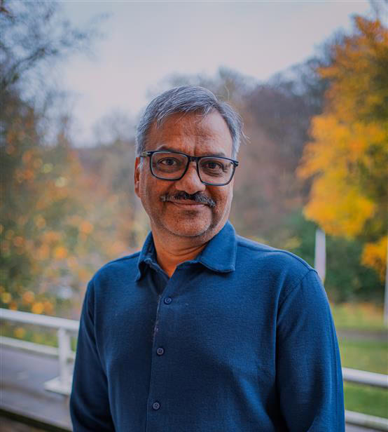 Een man met grijs haar en bril, lachend op een parkroute met herfstkleuren in de achtergrond.