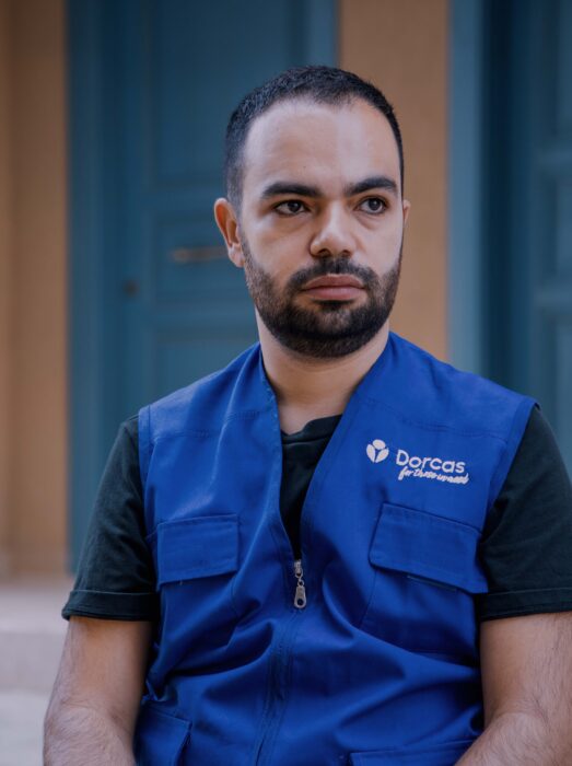Beeld van een werknemer van Dorcas in een blauw werkvest met logo, zittend en nadenkend aan de buitenkant van een gebouw.
