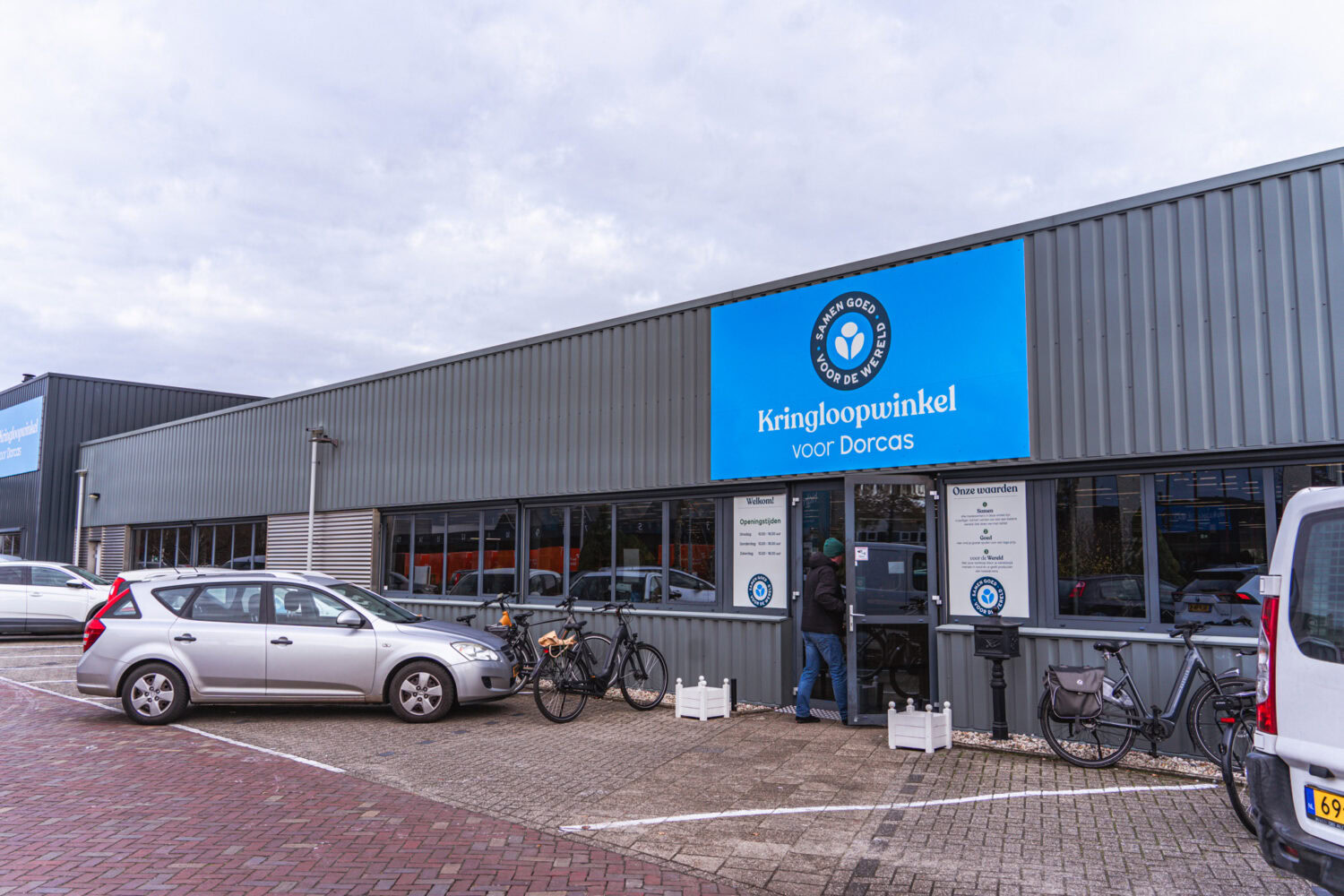 Kringloopwinkel voor Dorcas in een industrieel gebouw met parkeerplaatsen, fietsen en een blauwe bordgevel met logo en openingstijden, gericht op hergebruik en duurzaamheid.