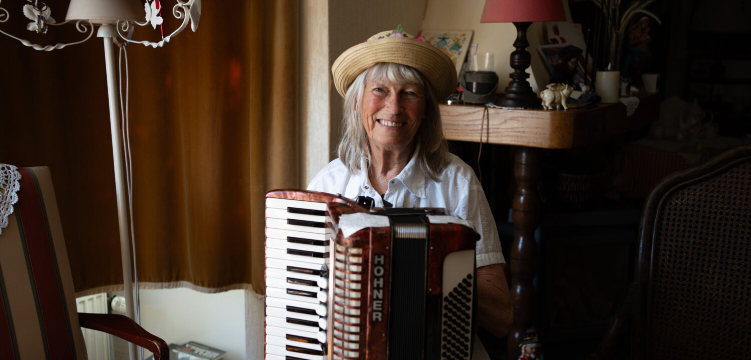 Een oudere vrouw met witte haren en een strooien hoed speelt op een accordeon in een gezellige woonkamer.