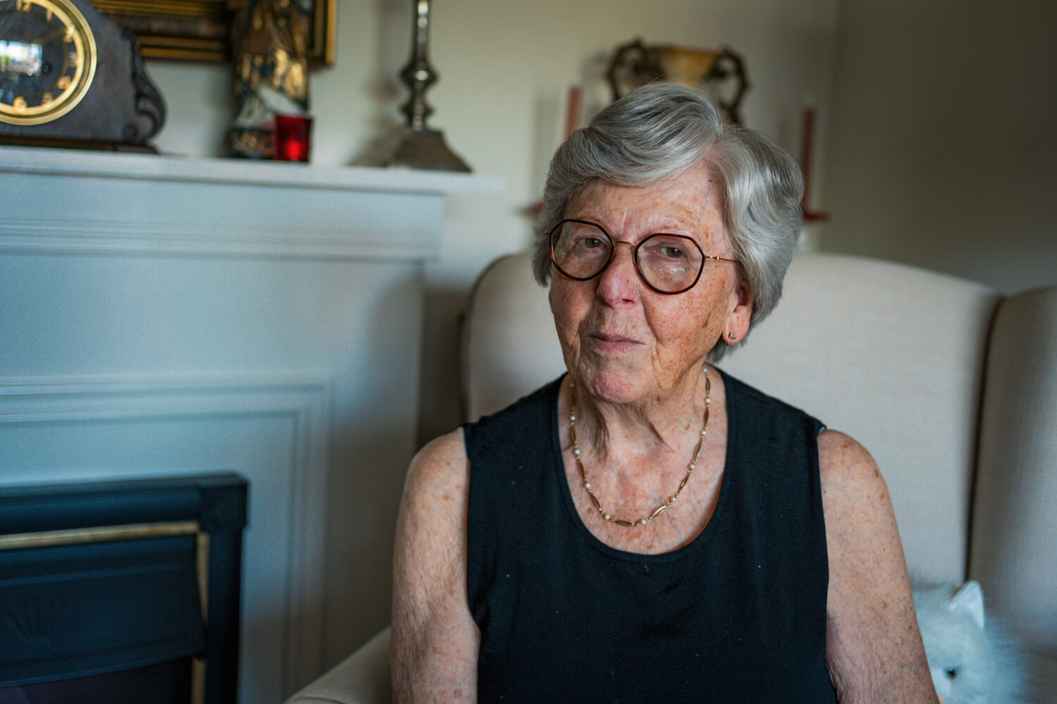 Zilverkleurige oudere vrouw met bril zit ontspannen in een woonkamer, draagt een zwart top en sieraden, sfeer van warmte en huiselijkheid.