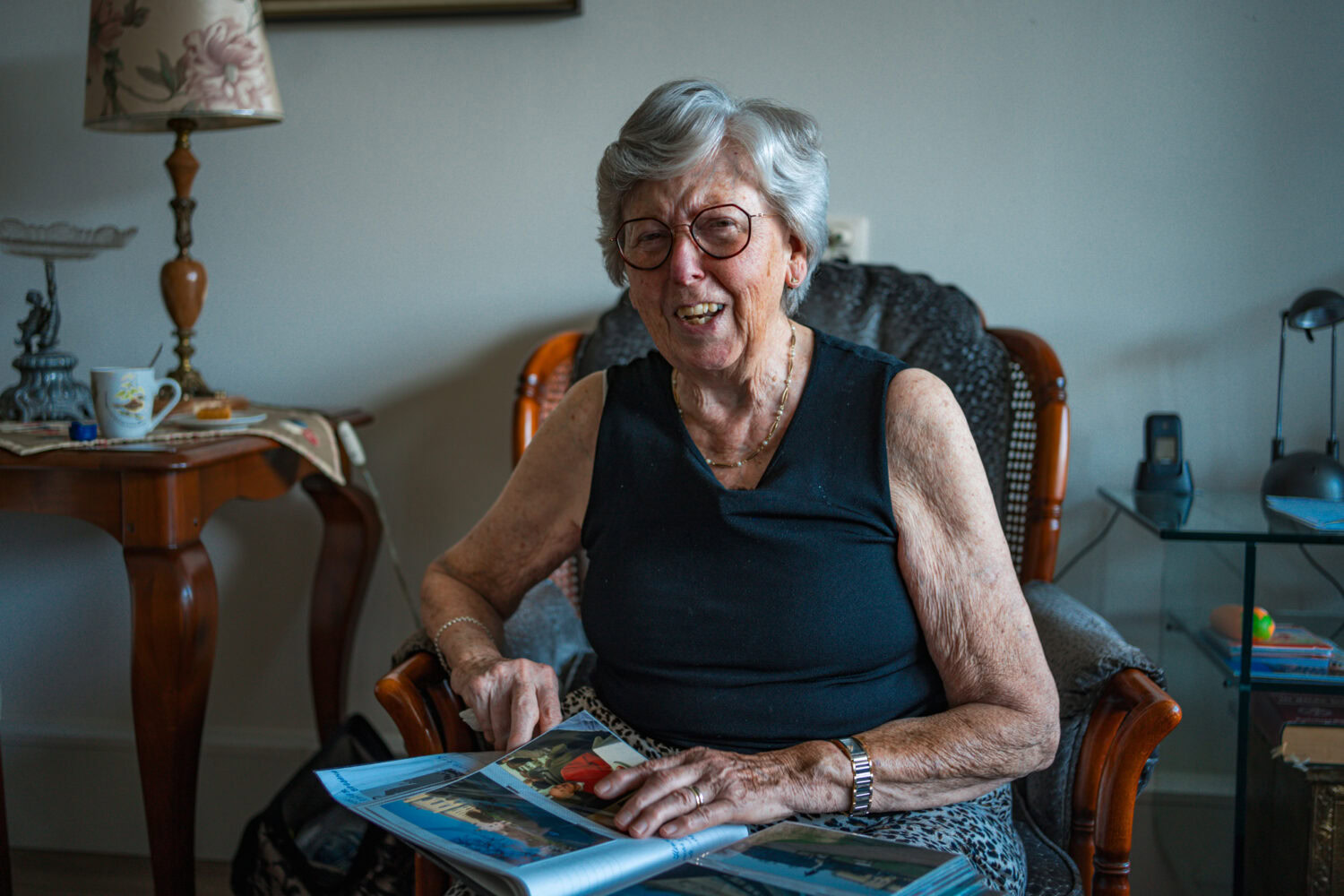 Oudere vrouw glimlachend in woonkamer, genietend van haar dag, met fotoalbum in haar hand, comfortabele stoel en gezellige interieur aan het achtergronds.