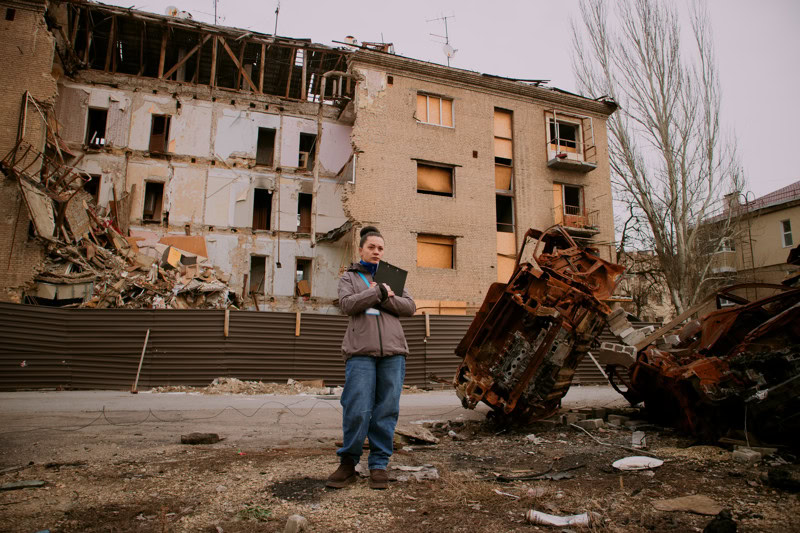 Een man staat met een tablet voor een verwoeste woning en auto, omgeven door schade en puin, illustrerend de impact van een ramp of incident op de omgeving.
