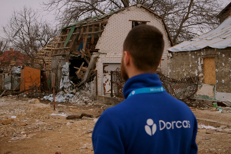 Een man met een Dorcas-jas kijkt naar een beschadigd huis dat is verwoest door een ramp, klaar voor herbouw en herstel.