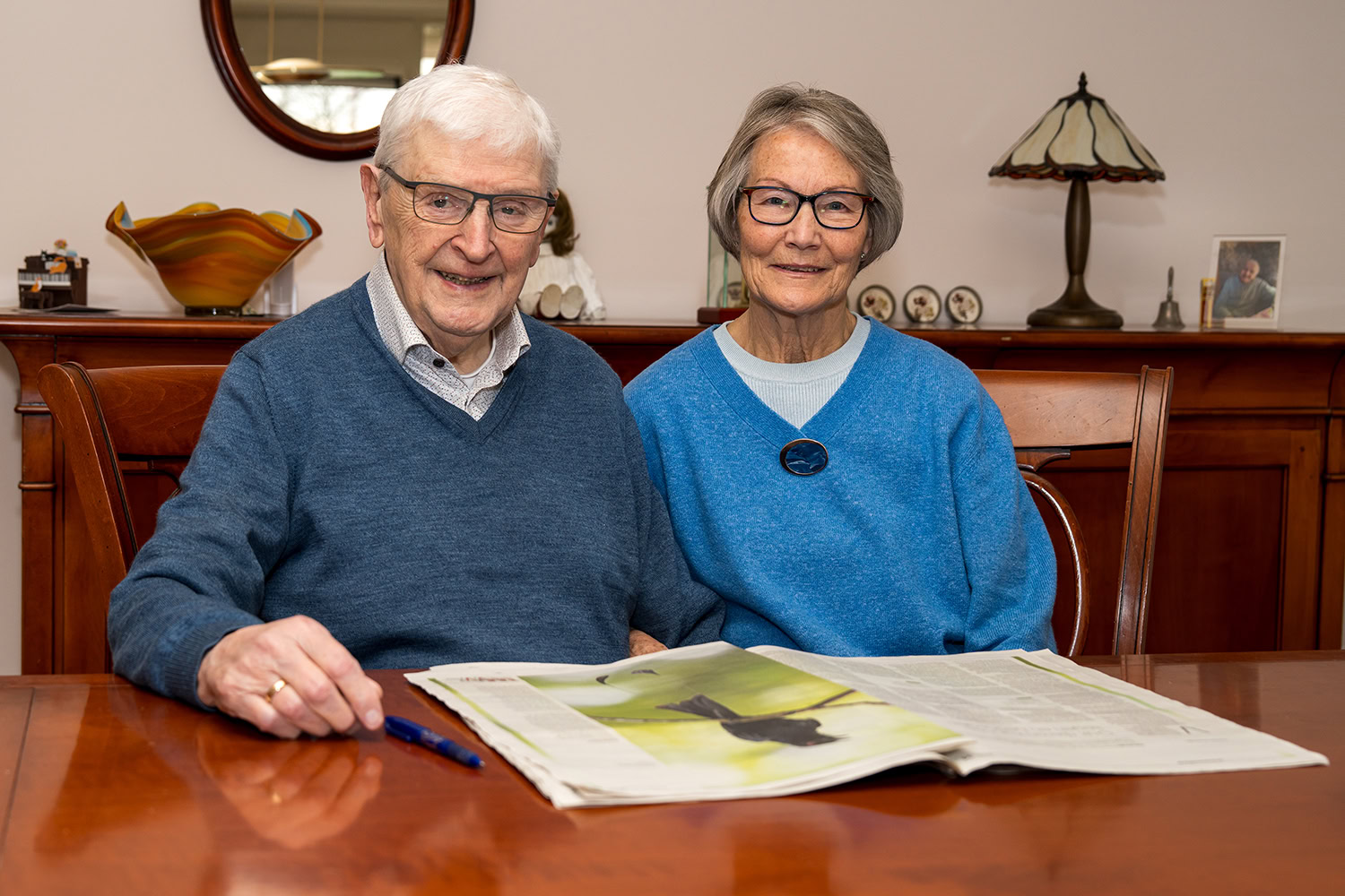 Een oudere man en vrouw glimlachen terwijl ze samen een krant bekijken, symboliserend de vreugde van geven en delen binnen de gemeenschap.
