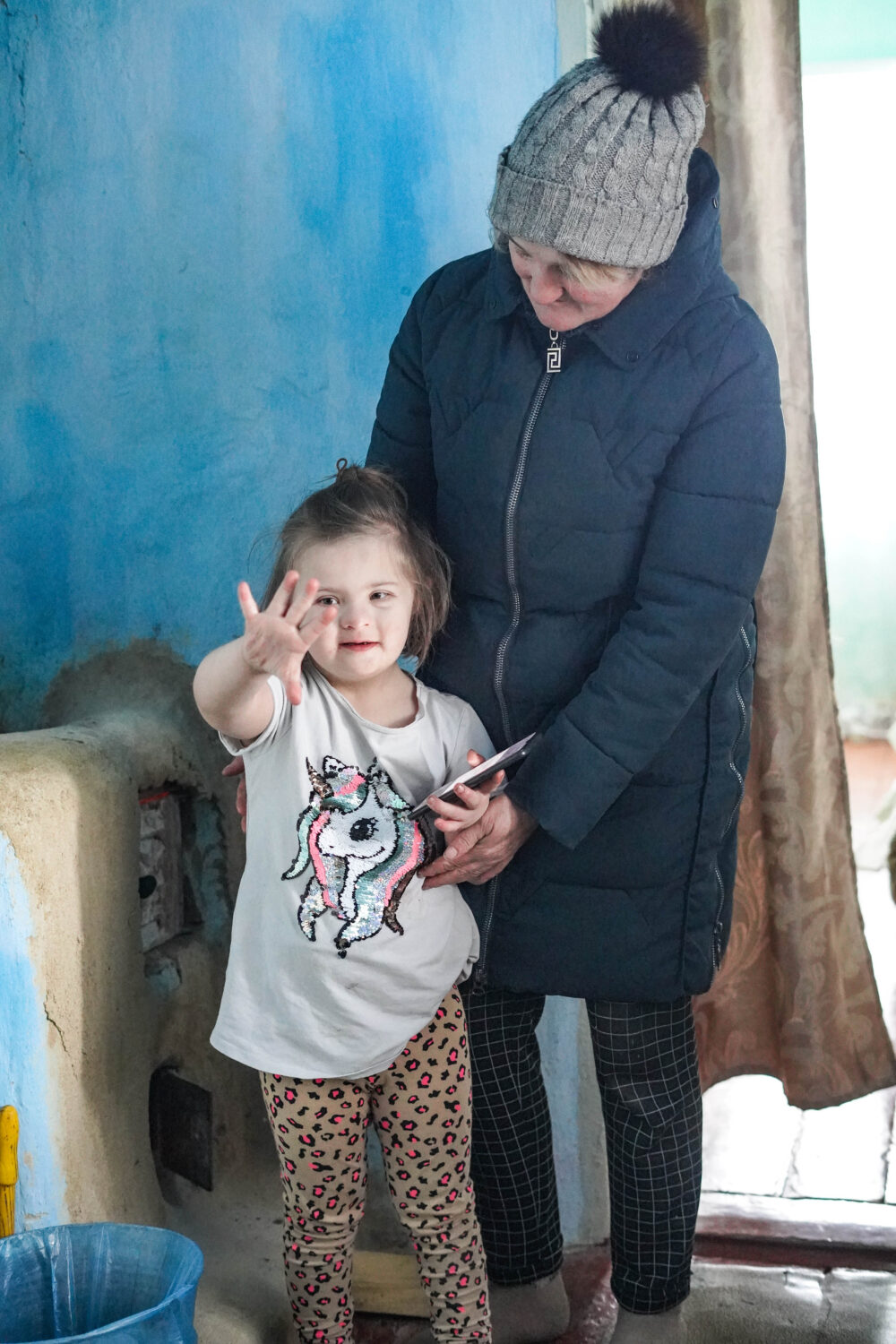 Een vrouw en een jong meisje in een huis in Oekraïne, waar de vrouw een mobiele verwarming vasthoudt terwijl het meisje vrolijk naar de camera wijst, illustrerend de behoefte aan warmte tijdens de win.