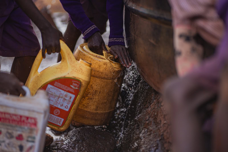 Een afbeelding van mensen die water uit een bron halen met behulp van een jerrycan en emmers, illustrerend duurzame watervoorziening en gemeenschapsinitiatieven.