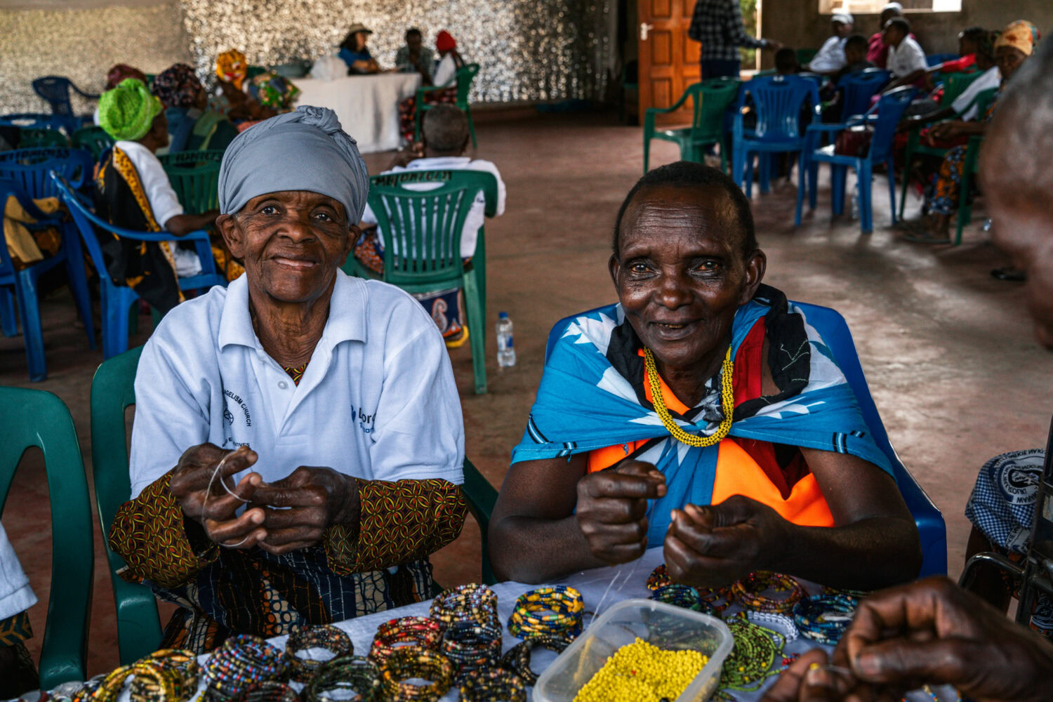 Twee vrouwen uit Tanzania lachen en genieten samen tijdens een activiteit, omringd door kleurrijke stoffen en een gemeenschap die samenkomt voor educatie en inspiratie.